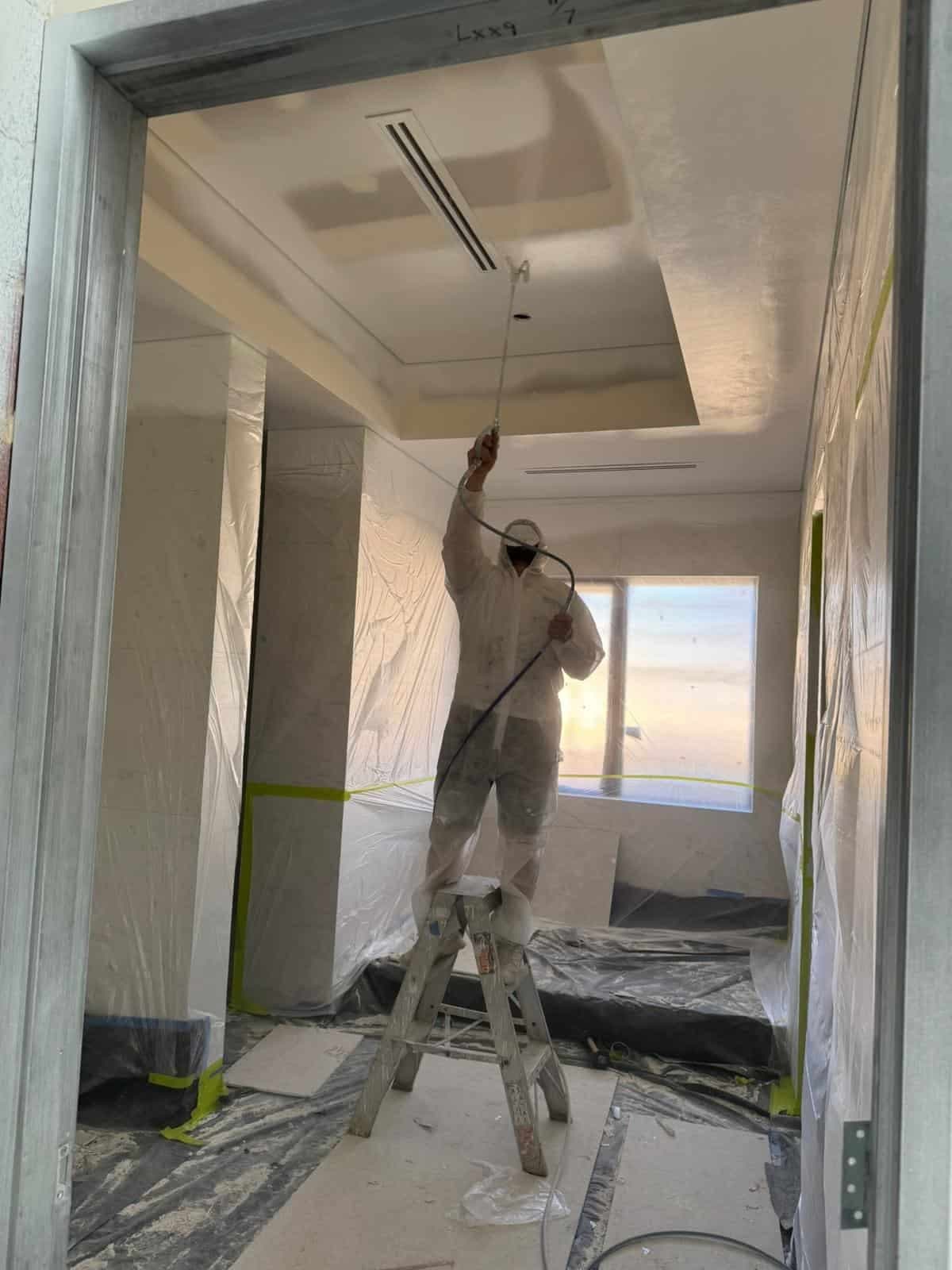 Painter in a white suit spray-painting a room with plastic-covered walls