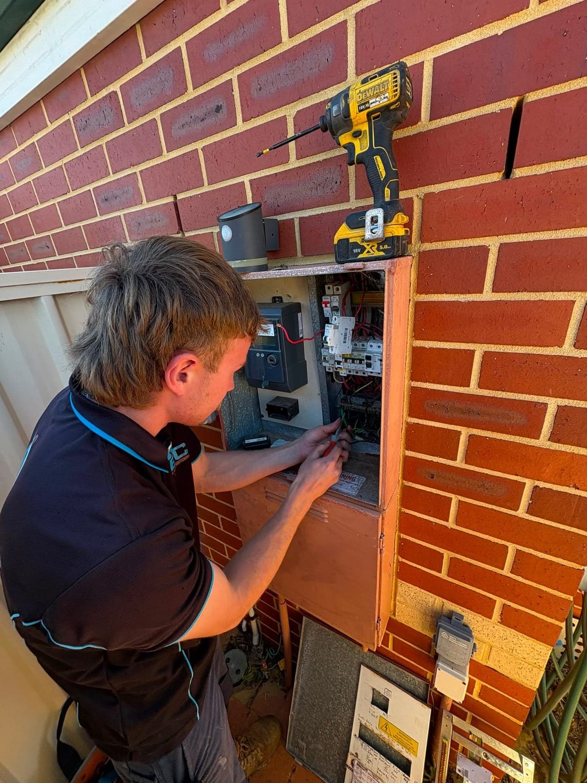 Electrician working inside a switchboard on a brick wall, drill parked on the brick