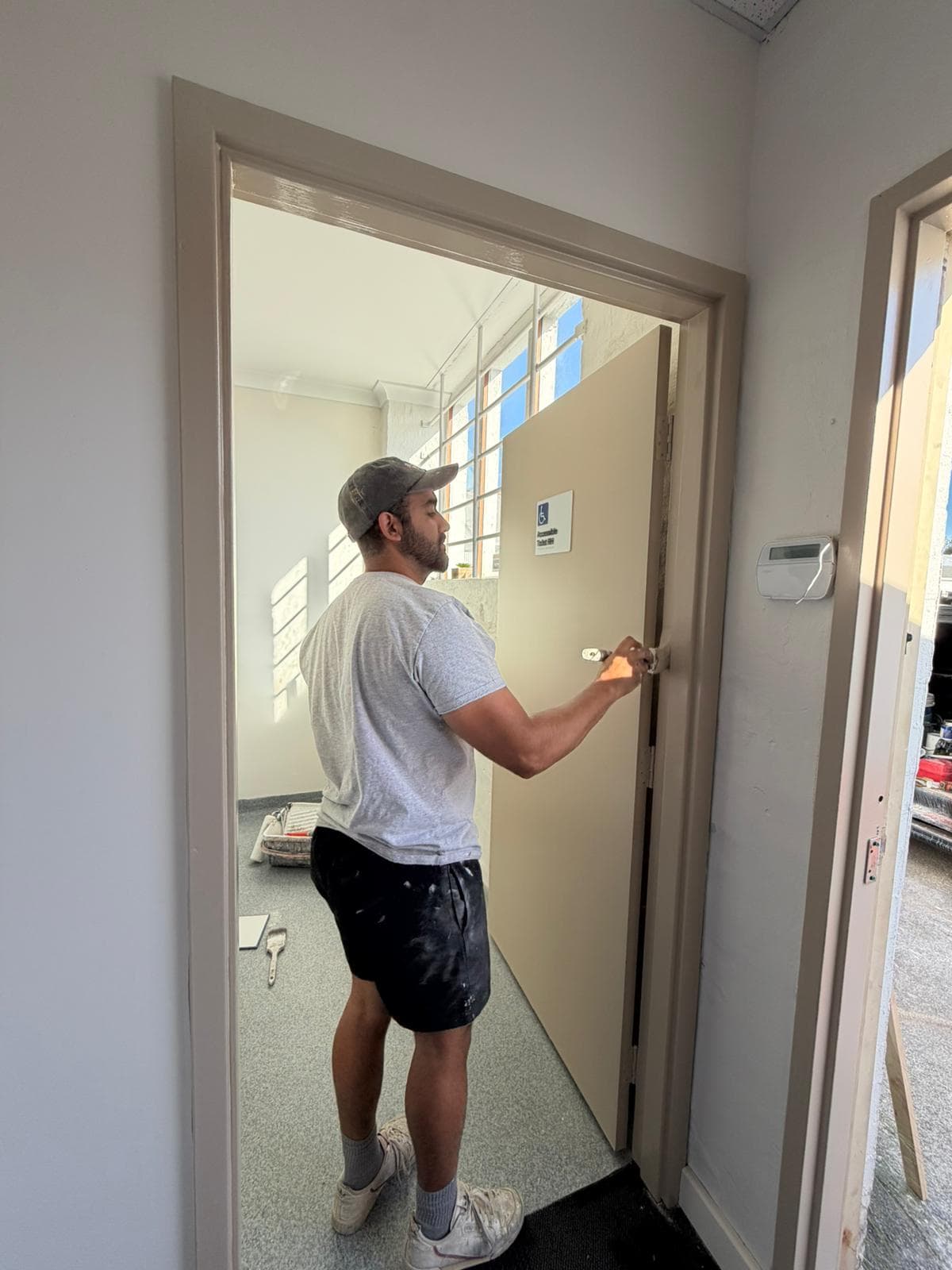 Daniel painting a door edge on a commercial site next to an accessible toilet sign, paint tray on the floor